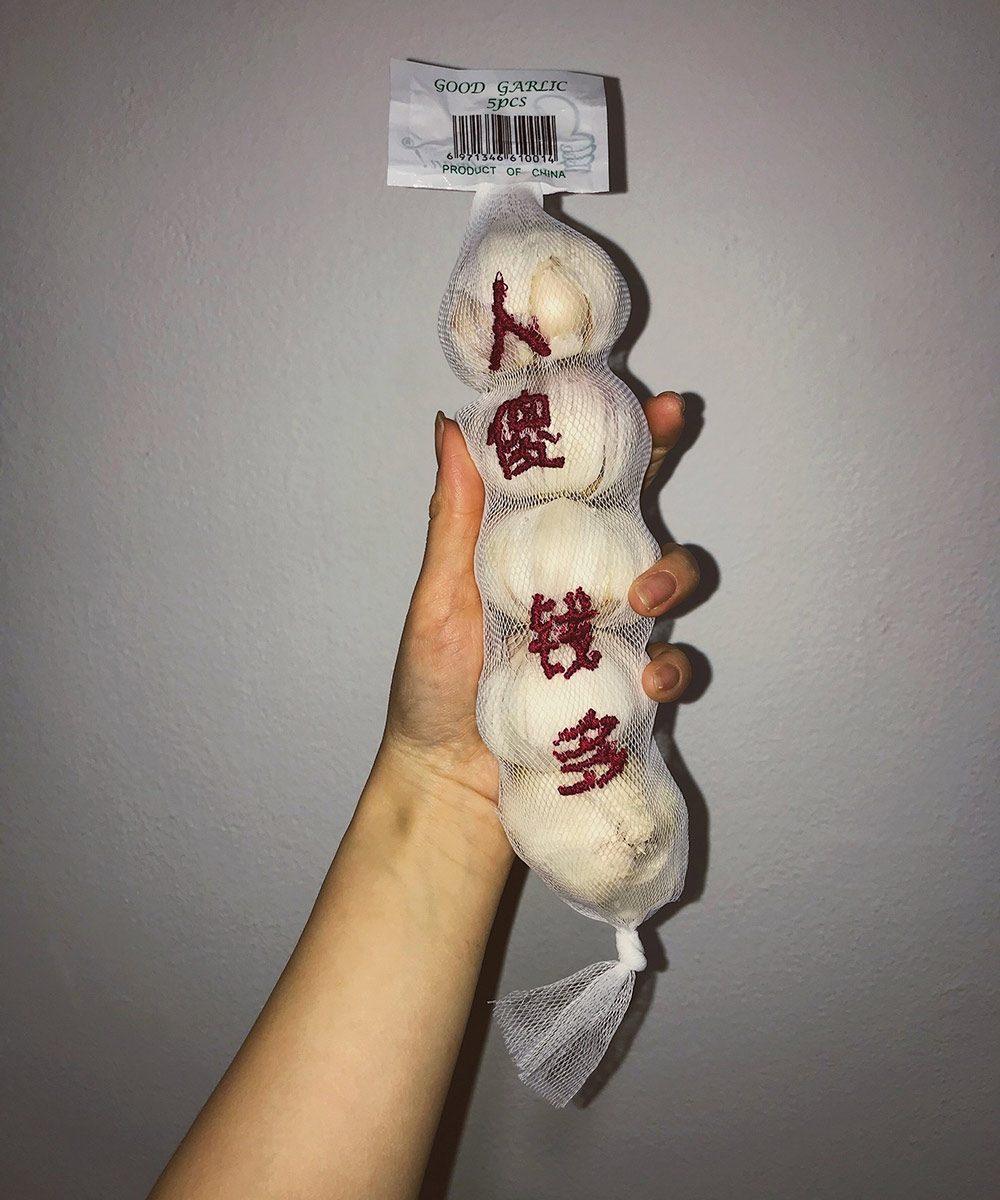 Close up on person's hand holding a mesh pacakge of white garlic bulbs with red characters running down the package.