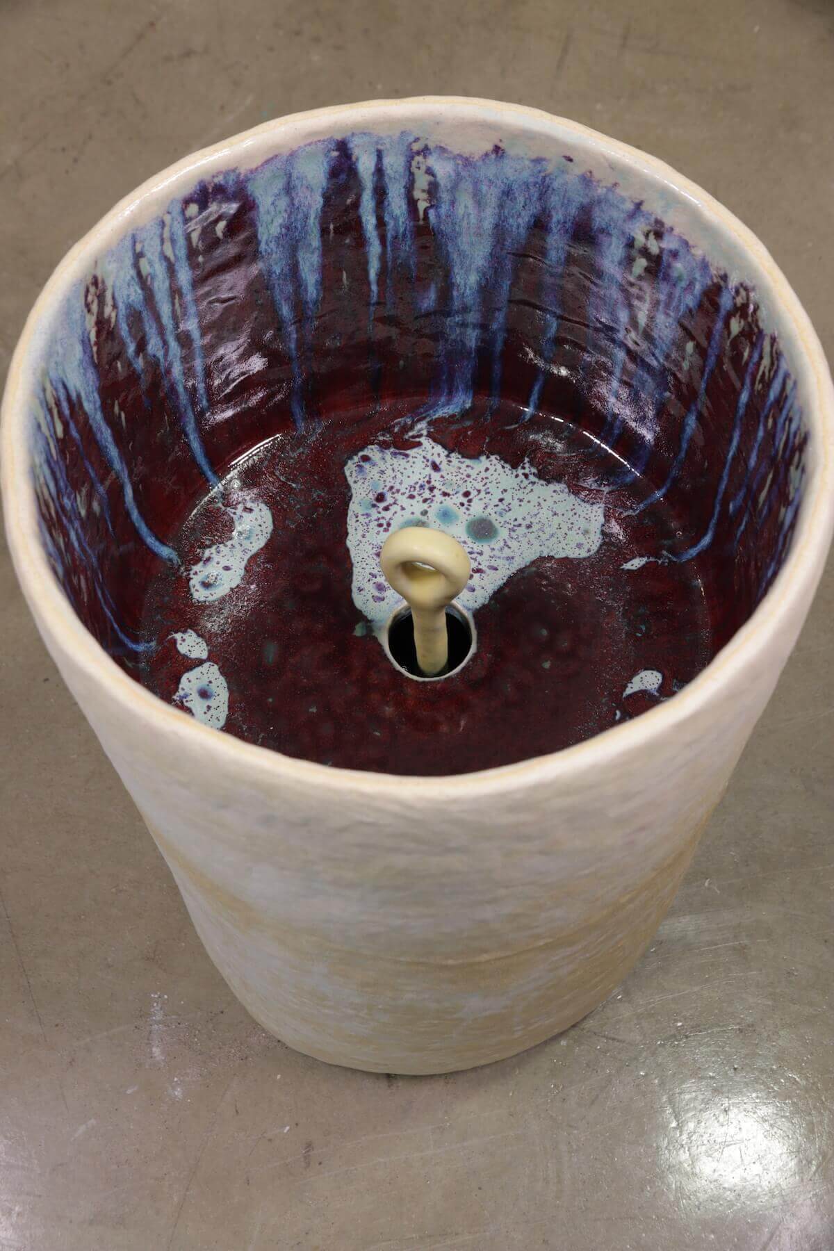 View into interior of white/cream ceramic cylinder with multicolored glaze dripping down sides to pool at the bottom of the structure.