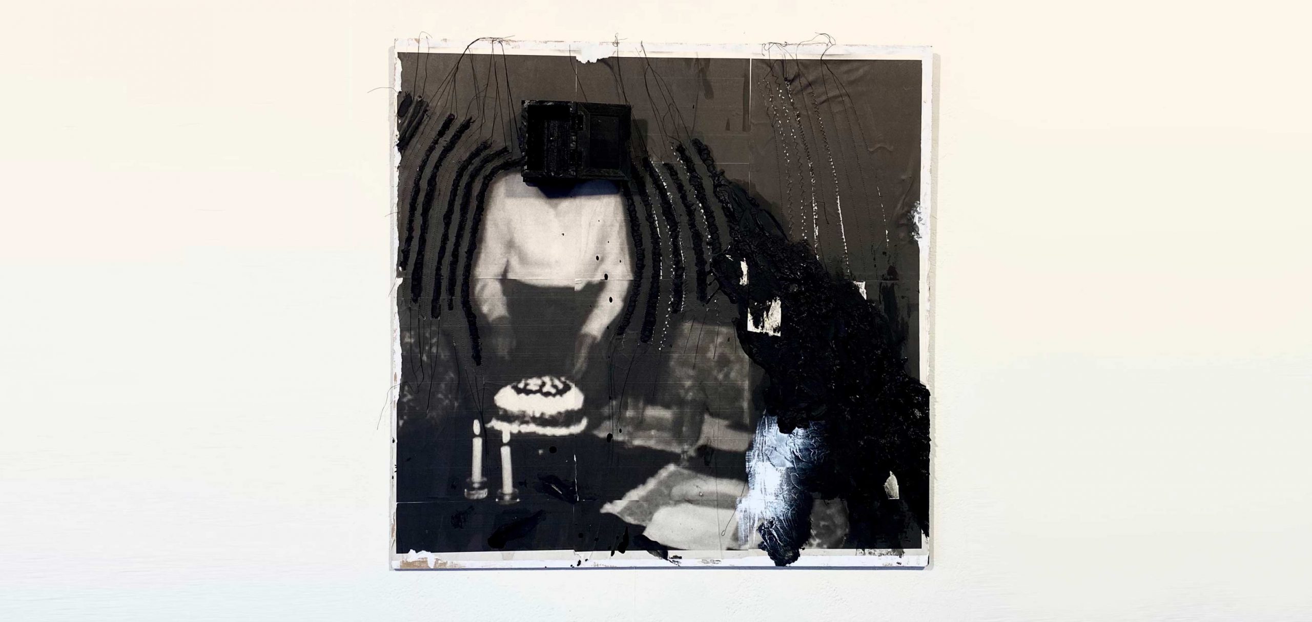 Black and white photo print altered with thread, acrylic paint, and wax. On the left is a person in white long-sleeve shirt with the head obscured by a black shape. The person is cutting a cake on a table with two candles and other semi-obscured table settings. The far right of the image is heavily obscured by added black ad white media..