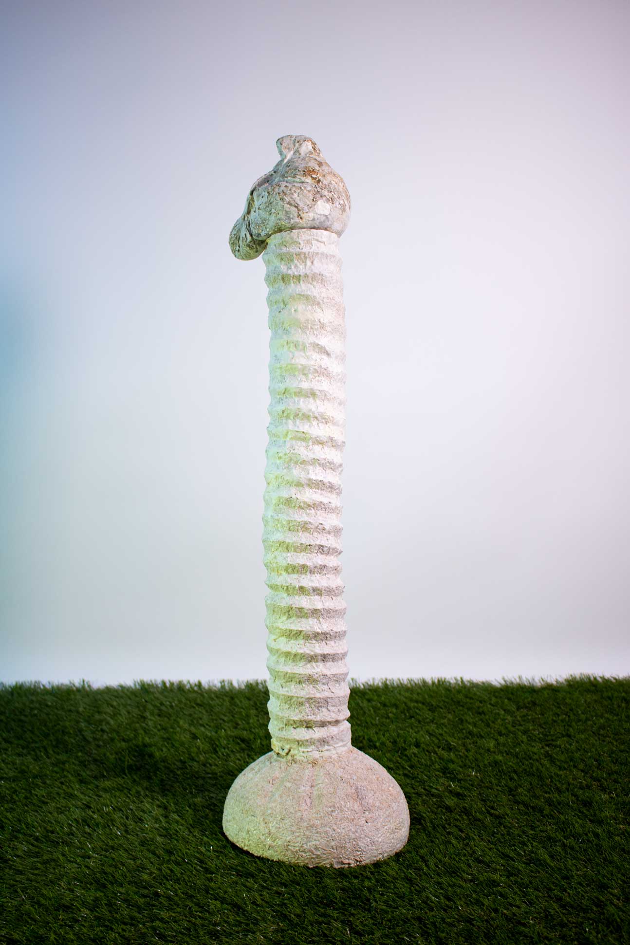 A white/cream textured, organic sculpture shaped like a post sits on faux grass in front of a white wall.