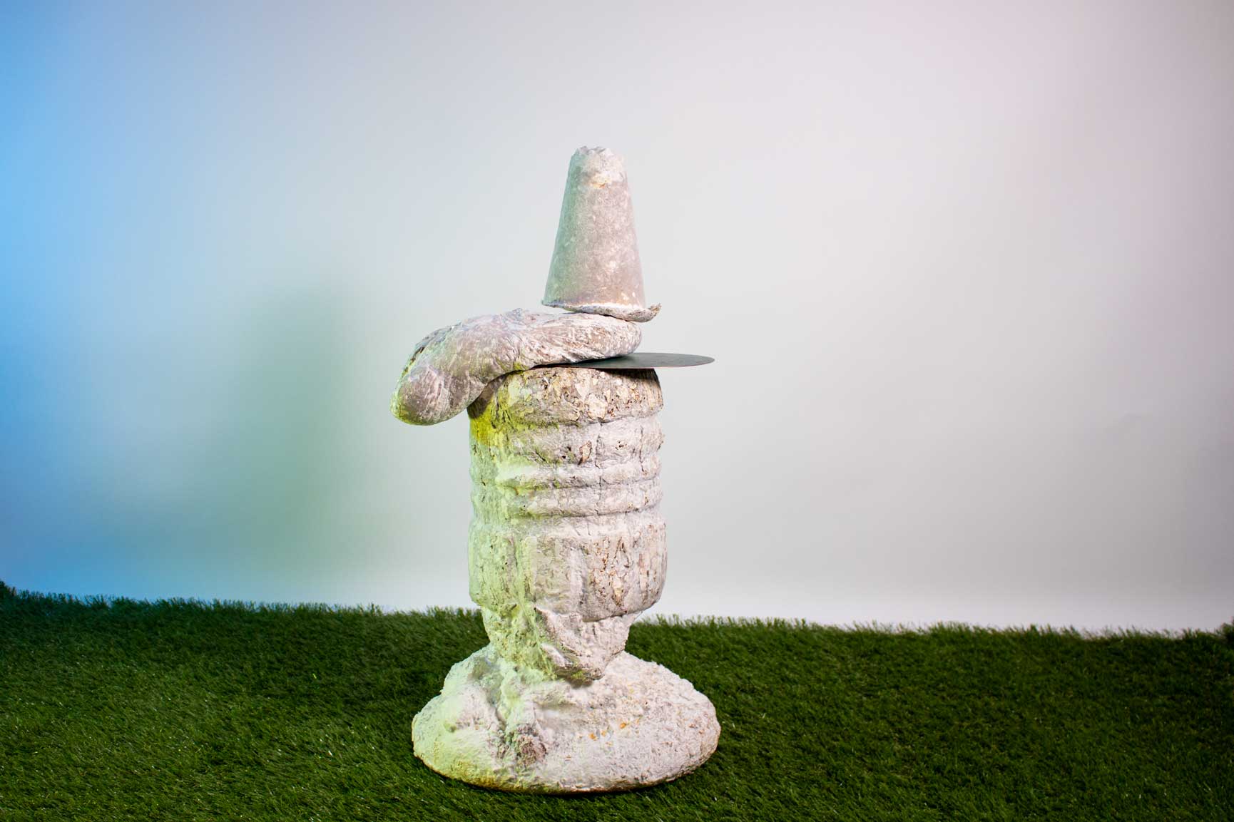 A white/cream textured, organic sculpture sits on faux grass in front of a white wall.