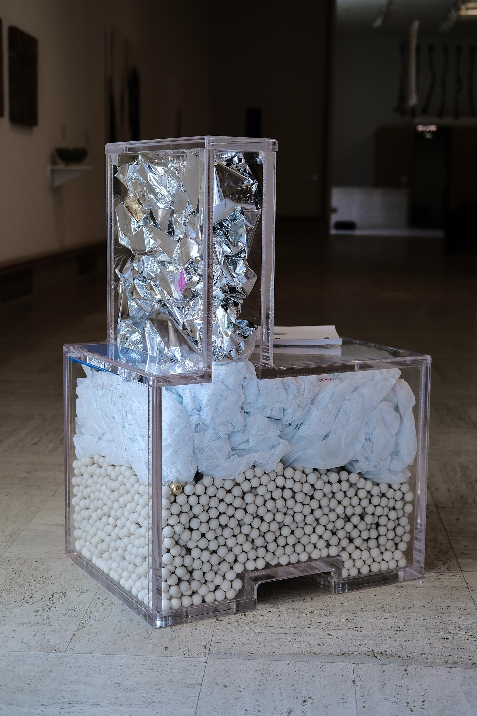 Rear view in darkened room of a ube chair made out of clear material filled with white fiber, ceramic balls, and silver foil.