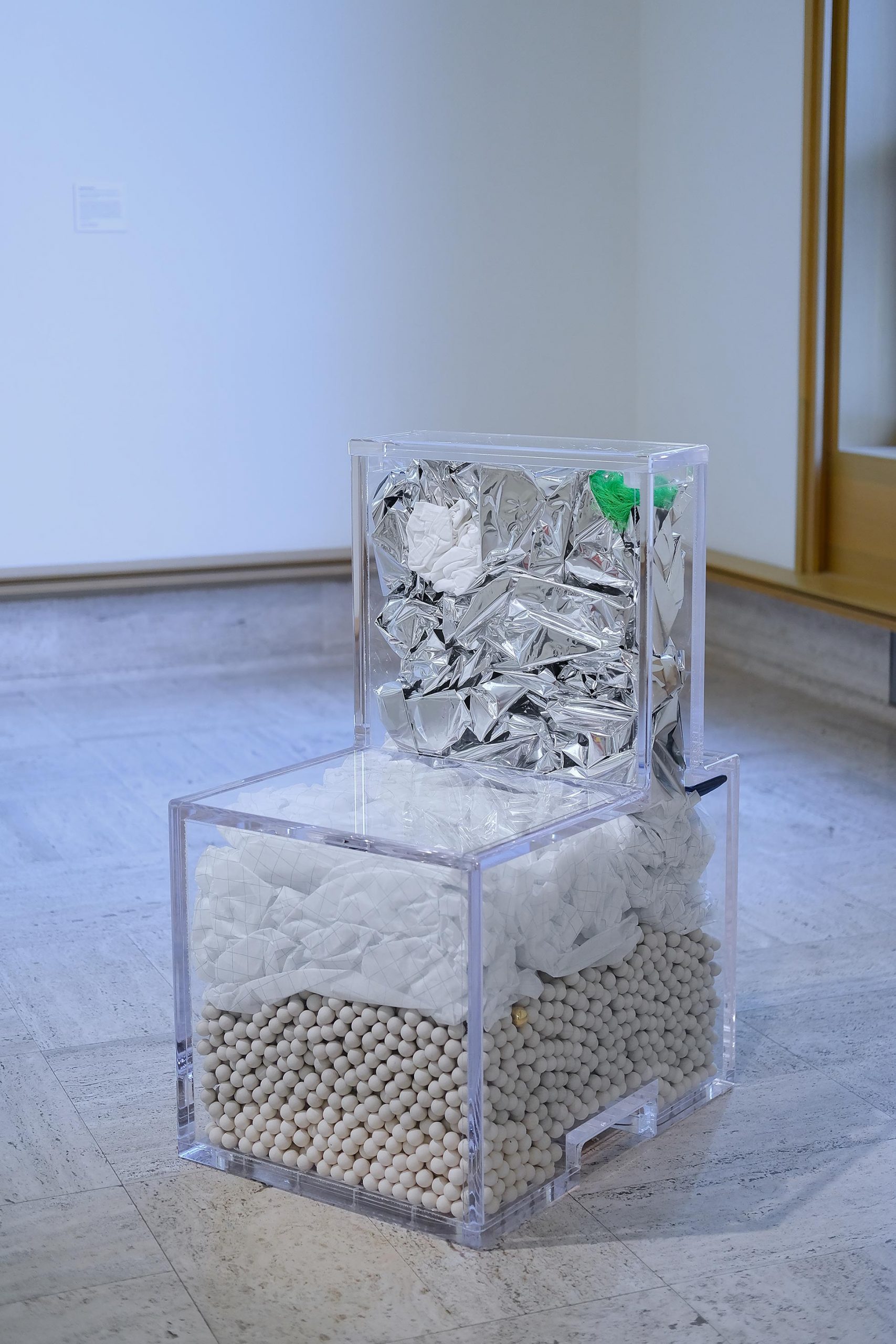 Cube chair made out of clear material filled with white fiber, ceramic balls, and silver foil.